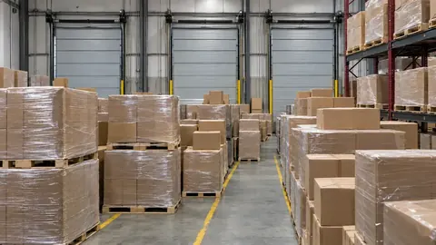 Warehouse loading dock with three closed doors, where the staging floor in front of them is filled with floor-stacked, shrink-wrapped pallets and cartons, leaving only a narrow path. Illustrative image.